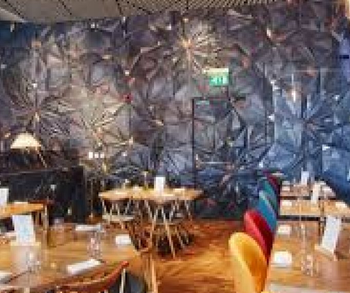 Dining area in front of a dark faceted feature wall with timber tables and bright upholstered chairs