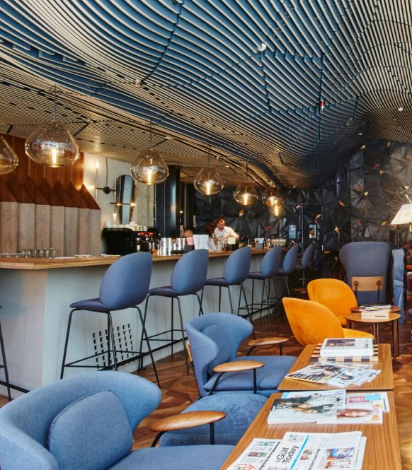 Coffee bar with blue stools under a curved slatted ceiling, lounge chairs and magazine tables