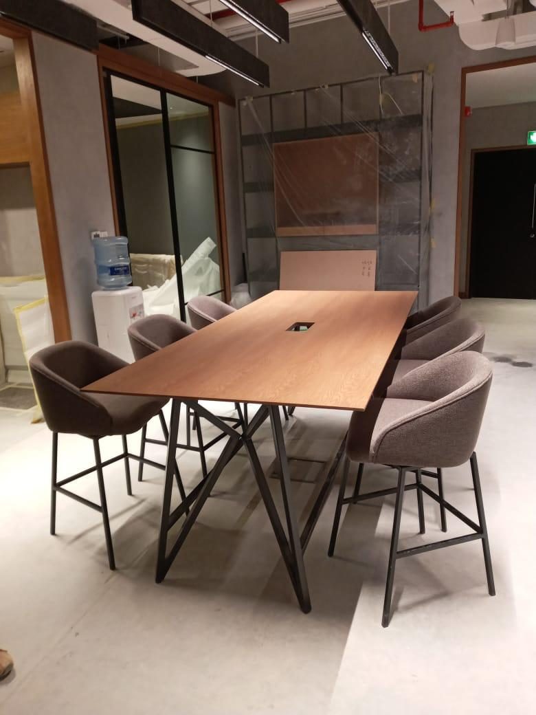 Timber meeting table with upholstered bar chairs beside black steel glazed partitions, during installation
