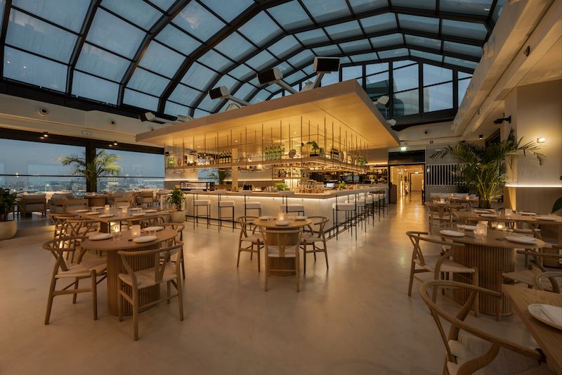 Glass-roofed rooftop restaurant with a central timber bar and oak dining chairs